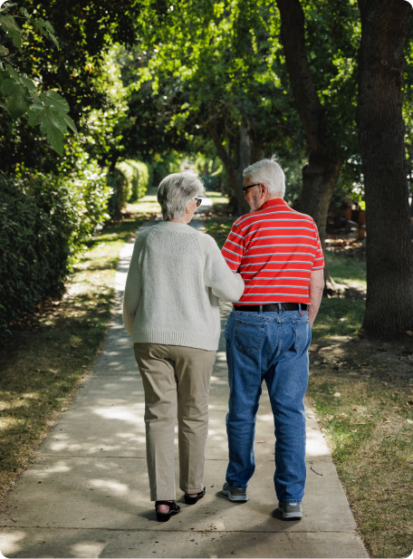 Two people walk down a tree-lined path, arm in arm, surrounded by greenery, enjoying a peaceful and sunny day.