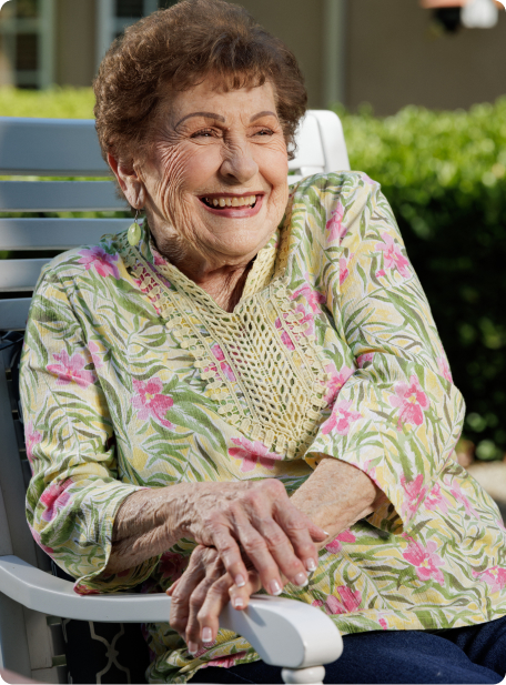 A smiling person with short hair sits outdoors in a floral shirt, enjoying a sunny day while leaning on a white chair.