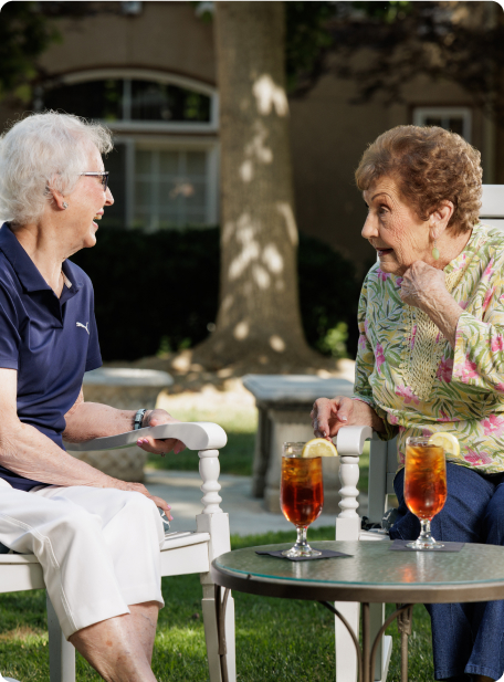 Two people are seated in a garden, engaged in conversation, with iced tea on the table. The background features a tree and building.