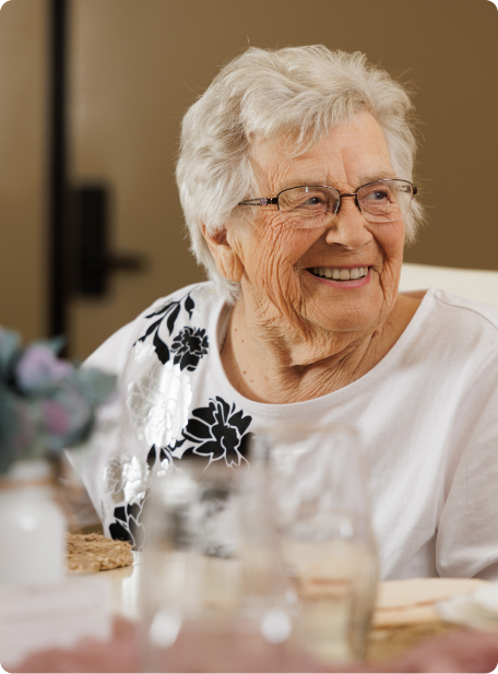 Person with glasses smiles, wearing a decorated white shirt. Blurred background suggests an indoor setting with soft lighting. No recognizable landmarks or buildings visible.