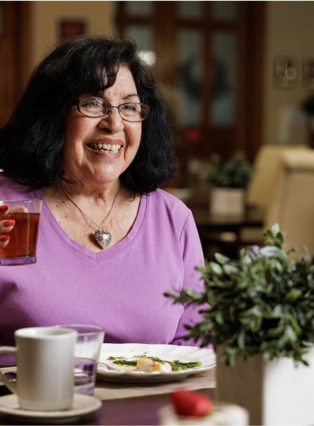 A person wearing glasses, in a purple shirt, smiling while holding a drink at a dining table with plants and tableware.