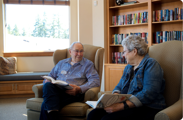 Two people are sitting in a cozy living room, reading books, surrounded by a bookshelf filled with colorful books. Large window view outside.