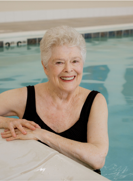 A person in a black swimsuit smiles, standing in a swimming pool, resting hands on the pool edge with water ripples visible.