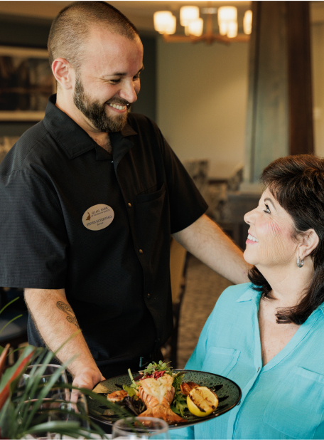 A person serves a meal to another person in a restaurant setting, showing friendly interaction. Indoor environment with warm lighting and decor.