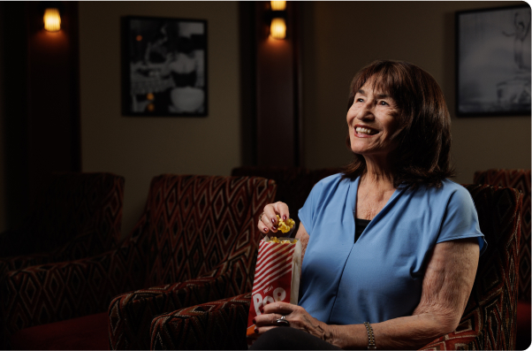 A person enjoys popcorn while sitting in a movie theater with patterned seats and dim lighting, smiling at the screen ahead.