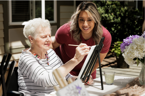 Two people enjoy painting outdoors on a sunny day, with one person helping the other. They are surrounded by flowers and art supplies.