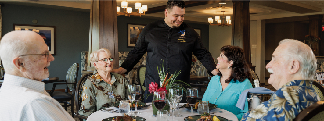 Four people sit around a table in a well-lit dining room with a person in a black chef coat smiling.