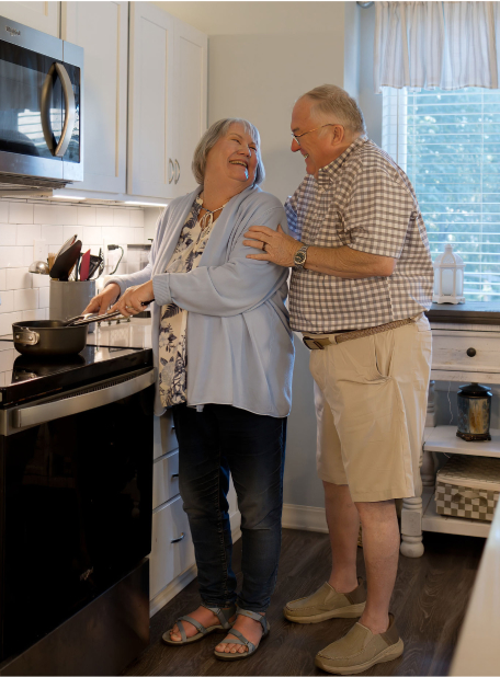 Two people smiling in a kitchen, one cooking at the stove. Bright natural light fills the space. Casual, cozy atmosphere.