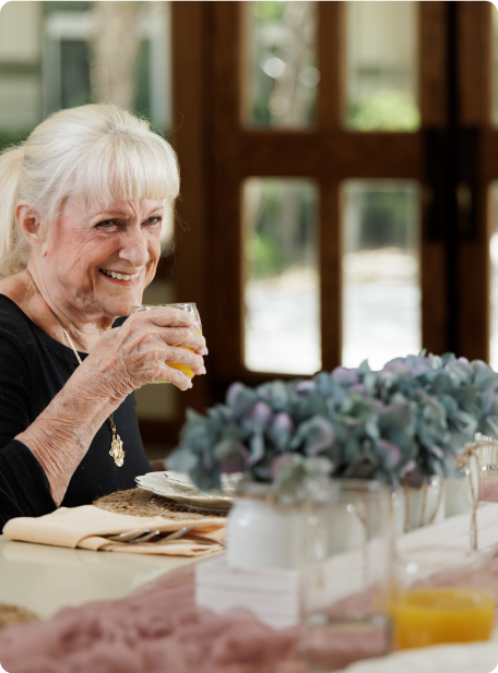 A person holding a drink sits at a table adorned with flowers, smiling warmly. Bright daylight filters through nearby windows.