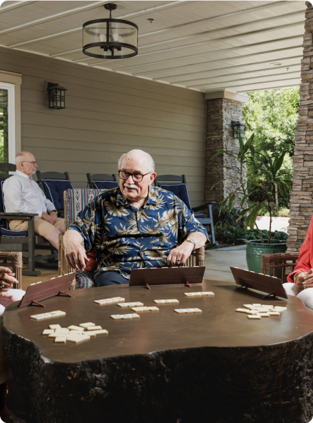 Four people are on a porch playing dominoes. One person is in a patterned shirt. The setting is relaxed and inviting, with greenery.