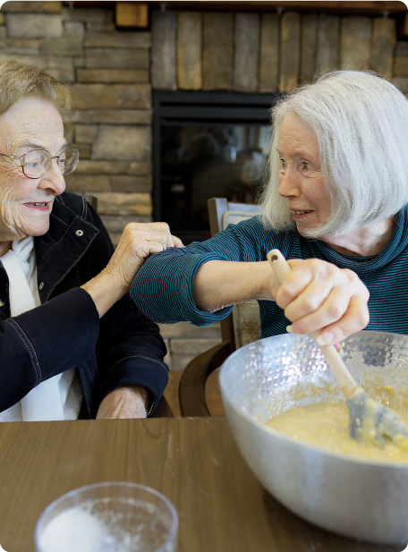 Two people sitting at a table, one mixing batter in a bowl. They are smiling and gently bumping elbows, with a fireplace behind them.