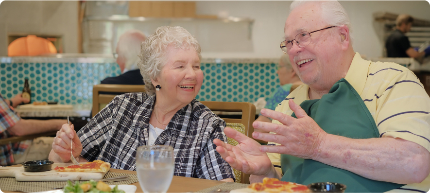 Two people are sitting at a restaurant table, enjoying food and conversation, with a cheerful atmosphere in the background.