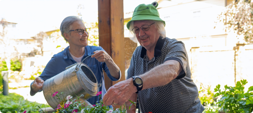 Two people smiling while gardening outdoors. One person waters flowers using a watering can. Bright sunlight and greenery create a lively atmosphere.