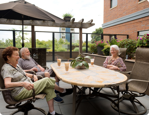 Three people sit around a table on a patio, enjoying drinks. The setting includes plants, an umbrella, and a brick building backdrop.