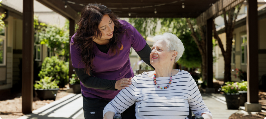 A person in purple assists another in a wheelchair outdoors, under a pergola, with garden views in the background.