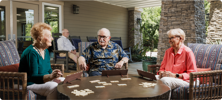 Three people sitting outside on patterned chairs, playing a board game on a round table. Stone and wooden walls in the background.