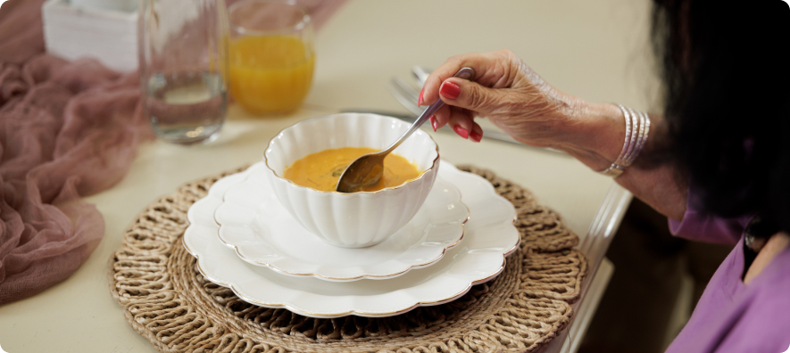 A person with painted nails enjoys a bowl of soup at a well-set table with a decorative placemat and drinks nearby.