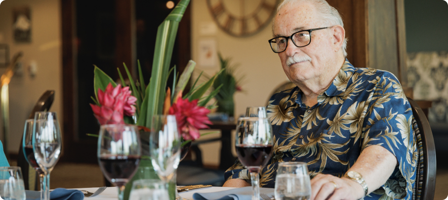 A person in a floral shirt is seated at a table set with wine glasses and tropical flowers indoors.