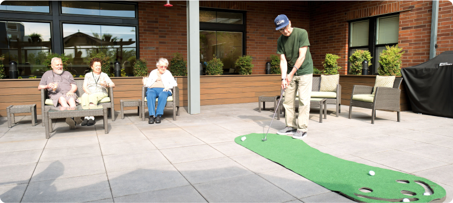 Four people are enjoying a game of mini-golf on a sunny patio, surrounded by chairs and greenery, outside a brick building.