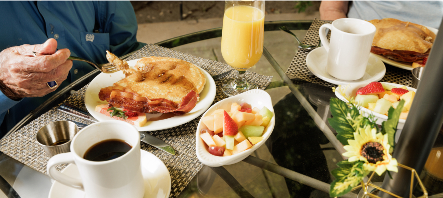 Two people enjoying breakfast at a glass table with coffee, orange juice, pancakes, bacon, and fruit salad, in an outdoor setting.