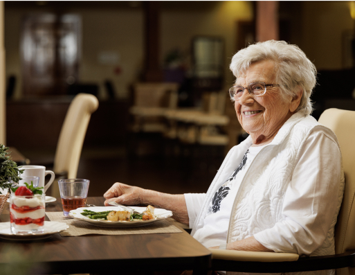 A person is seated at a dining table, smiling, with food and drink. The background shows a cozy, warmly-lit room.