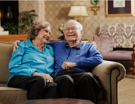 Two people sitting on a sofa smiling in a cozy, warmly lit living room with patterned decor and a table lamp in the background.