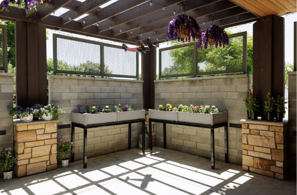 A sunlit garden area with raised planter boxes, stone pillars, and hanging flowers under a pergola, surrounded by cement block walls and greenery.