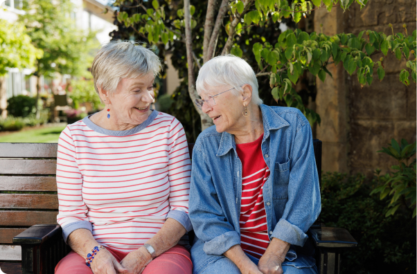 Two people smiling on a bench, surrounded by green trees and shrubs in a sunny outdoor setting. They appear to be engaged in conversation.