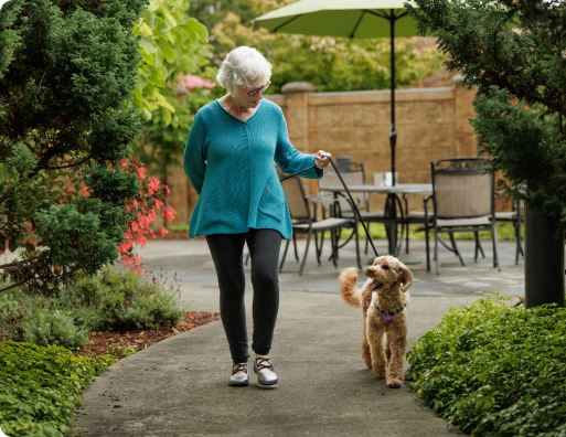 A person walks a dog along a garden path, surrounded by greenery and patio seating, under an umbrella.