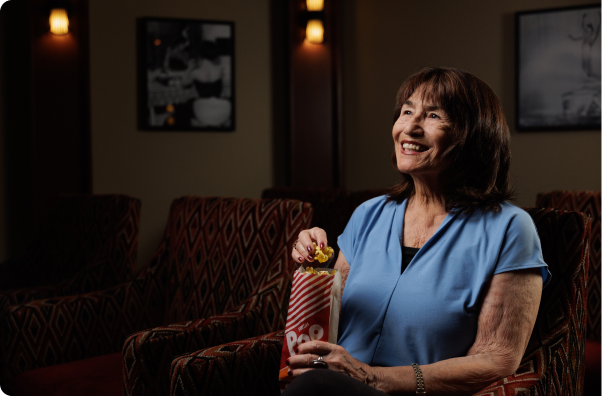 A person in a blue shirt enjoys popcorn while seated in a dimly-lit theater with patterned chairs and framed pictures on the wall.