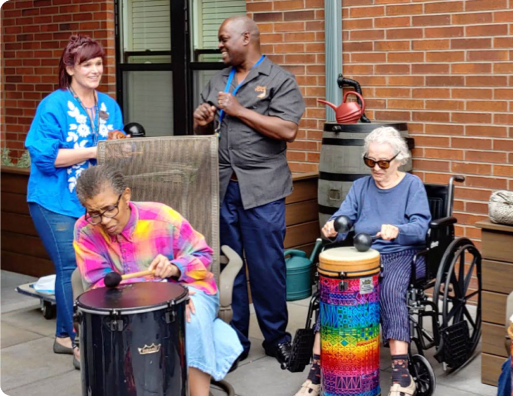 Three people play colorful percussion drums outdoors, while two others stand nearby observing and smiling. The setting is casual and engaging.