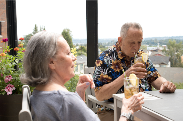 Two people sitting on a patio enjoying drinks. The scene includes flowers and a hilly background landscape, creating a relaxed atmosphere.