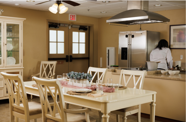 A person in a kitchen prepares food beside a dining table set for four, with a modern refrigerator and decorative flowers.