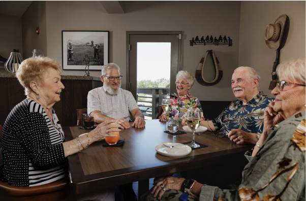 Five people sit around a table enjoying drinks in a cozy room; wall art and decorations create a warm, inviting atmosphere.