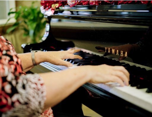 A person wearing a patterned shirt plays a Yamaha piano, with focus on hands. A plant is visible in the background.