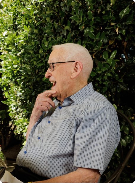 A person smiles while sitting in a chair, surrounded by lush green foliage on a sunny day. The setting is outdoors.