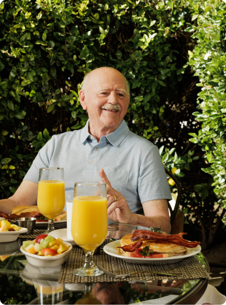 A person sits at an outdoor table with breakfast food, including pancakes and orange juice, against a backdrop of leafy greenery.