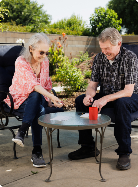 Two people sit outdoors at a round table playing a game, surrounded by plants and a low brick wall in the background.