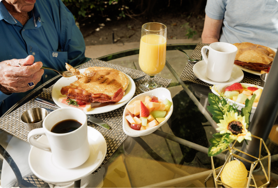 Two people are enjoying breakfast outdoors. The table features pancakes, bacon, fresh fruit, coffee, and orange juice, with a small floral decoration.