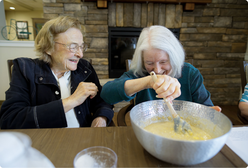 Two people sharing a joyful moment while mixing batter in a large bowl, sitting indoors beside a stone fireplace.