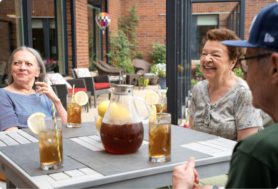 Three people enjoying iced tea on a sunny patio with a brick wall backdrop and outdoor seating. A decorative hot air balloon visible.