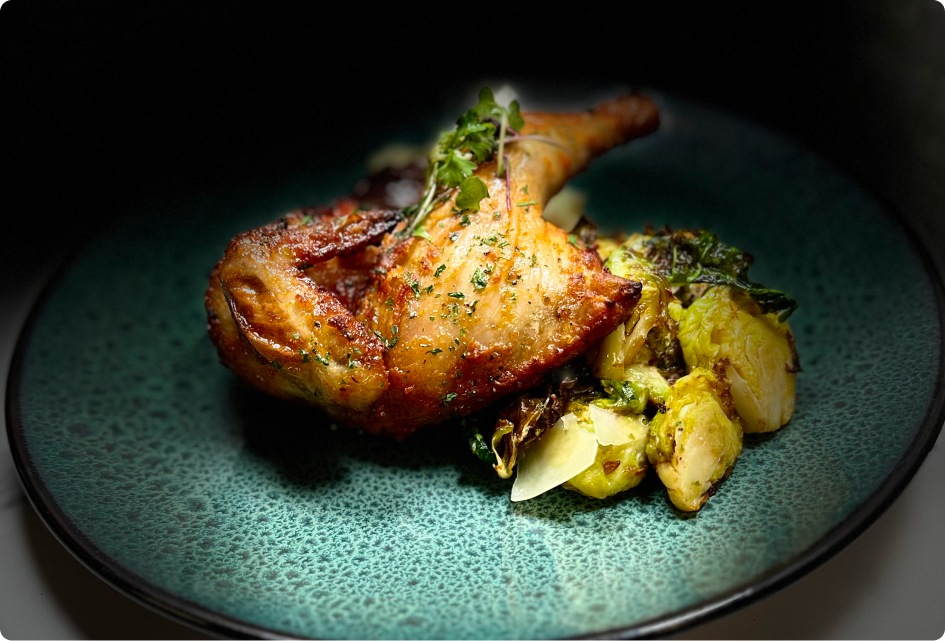 Roasted chicken leg garnished with herbs, served on a speckled green plate alongside sautéed Brussels sprouts. Appealing and vibrant culinary presentation.