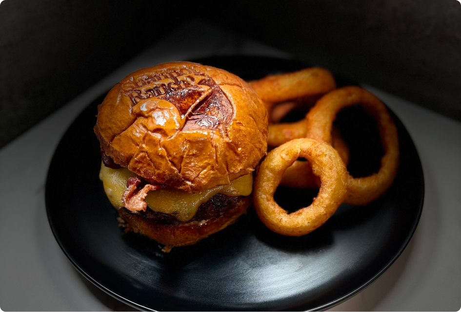 A cheeseburger with bacon on a branded bun is served alongside onion rings on a black plate in dim lighting.