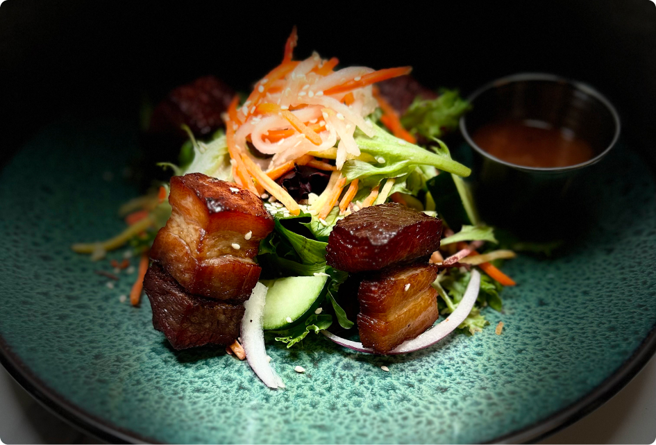 A vibrant salad with sliced vegetables, glazed pork cubes, and a side of sauce on a textured green plate, artfully presented.