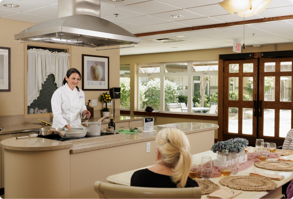 A person in a chef's uniform prepares food in a modern kitchen while another person watches, sitting at a table set for dining.