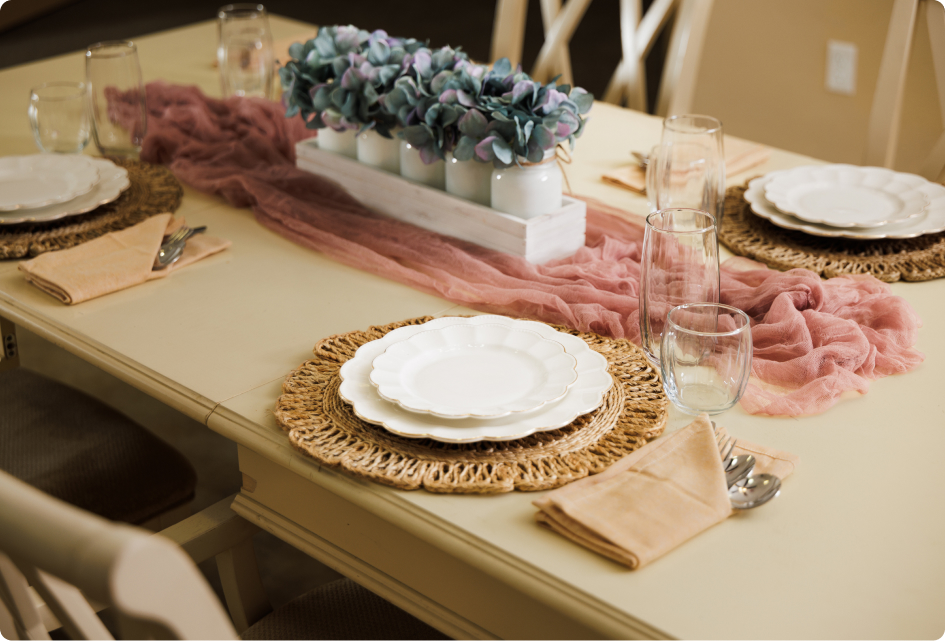 A neatly arranged dining table with woven mats, white plates, pink runner, clear glasses, and a floral centerpiece. Cozy and elegant setting.