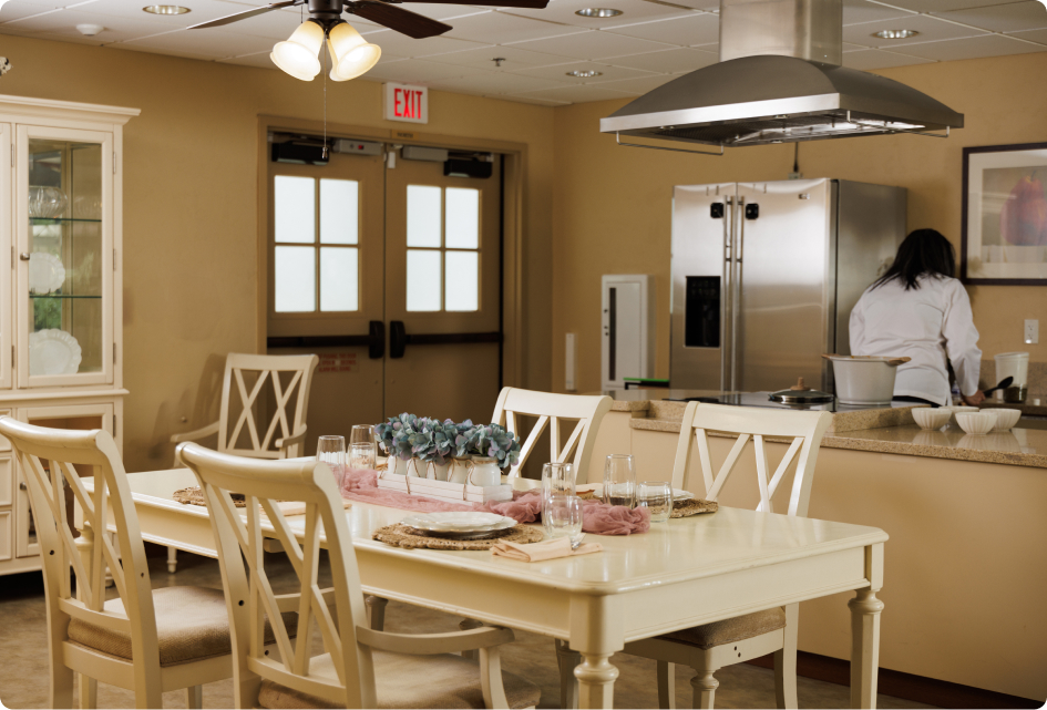 A kitchen with a dining table set for four. A person is cooking, and modern appliances are in the background.