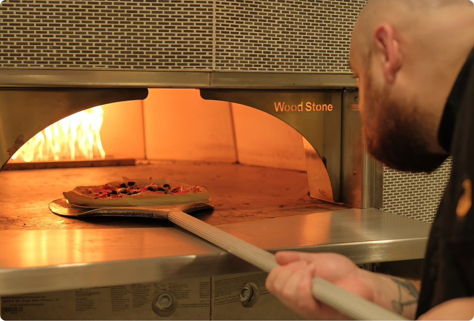 A person uses a peel to place a pizza into a Wood Stone oven with visible flames inside.