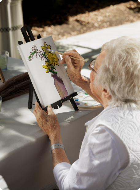 A person is painting a colorful flower bouquet on a small canvas set on an easel, outdoors in sunlight.