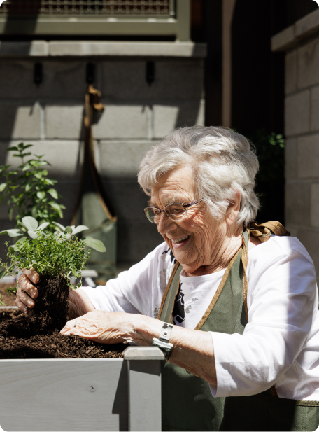 A person with white hair gardening, smiling while holding a small plant. The background includes a brick wall and gardening tools hanging.